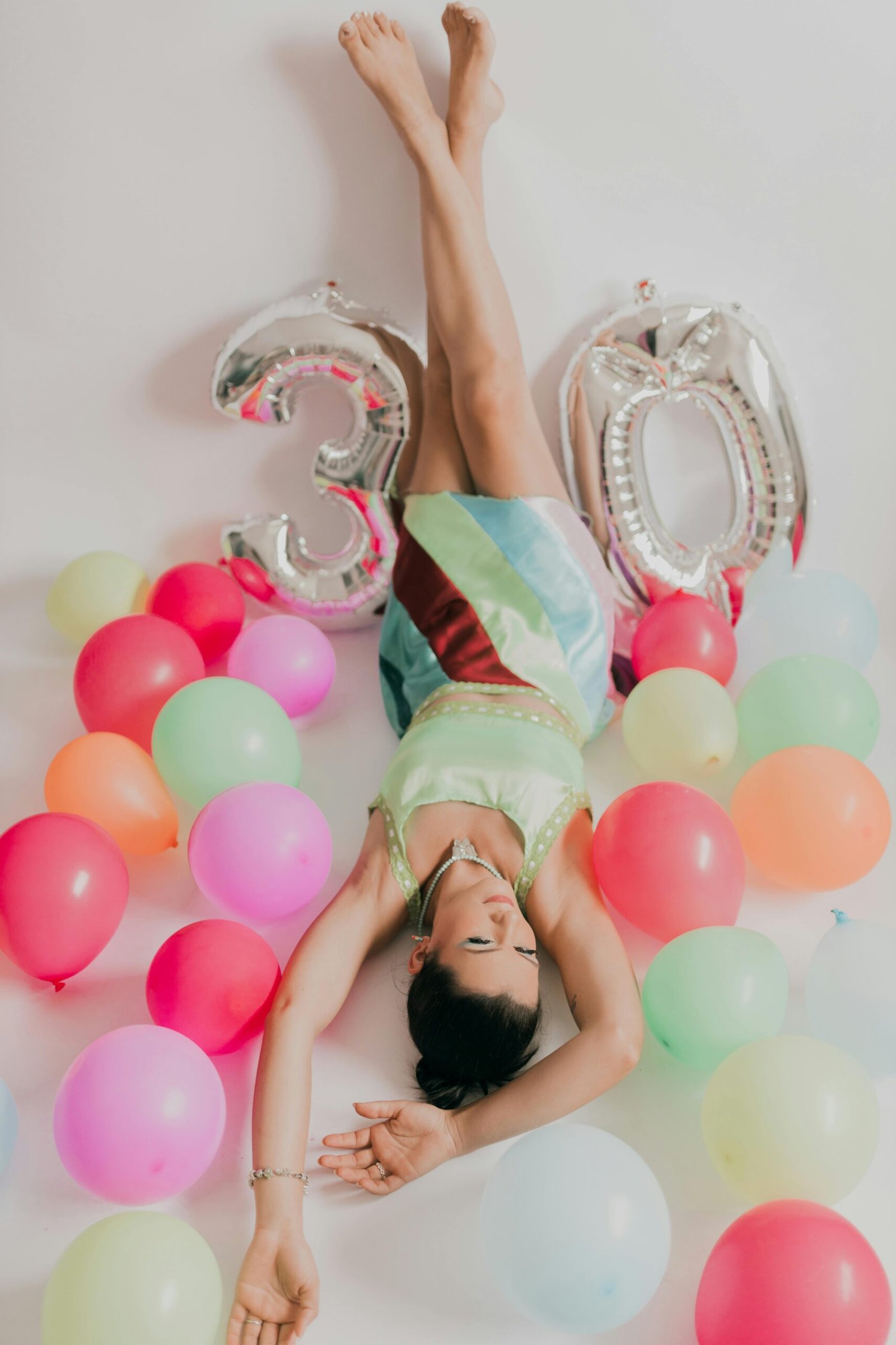Crise de la trentaine : entre fête et questionnements Femme allongée entourée de ballons colorés avec des ballons argentés en forme de “30”, symbolisant la crise et la fête des 30 ans.